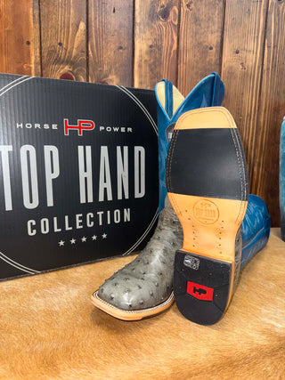 A pair of HP Power Top Hand collection cowboy boots displayed on a tan hide rug in front of a wooden wall. One boot is shown from the side in blue leather, and the other is turned to show the sole, which features a black and tan leather heel with a red HP logo.