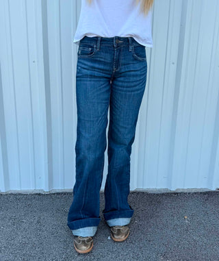 A person wearing dark blue flare jeans with a white t-shirt and brown shoes, standing in front of a white corrugated metal wall.