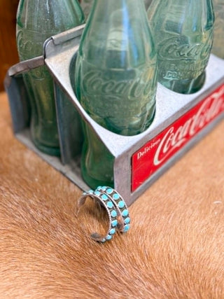 Set of green Coca-Cola bottles in a metal carrier with a turquoise earring on a wooden surface.