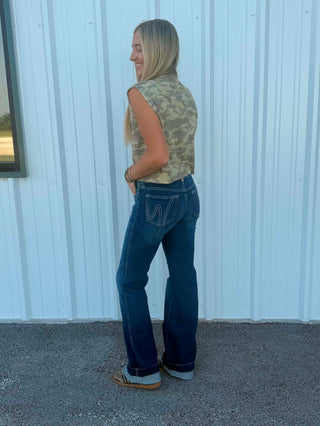 A woman stands in profile outdoors against a white corrugated metal wall, wearing a sleeveless camouflage top, dark blue jeans, and brown shoes.