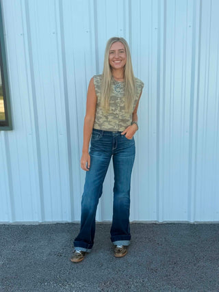 A woman stands outdoors in front of a white corrugated metal wall, wearing a sleeveless camouflage-patterned top, blue jeans, and brown shoes.