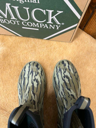 A pair of Muck Boot Company rubber boots with a green and gray tiger-stripe camouflage pattern, shown next to their branded box on a tan textured surface.