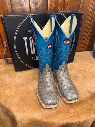 A pair of turquoise blue and gray ostrich-textured cowboy boots displayed in front of a black box with a circular logo, set on a brown fur surface against a wood-paneled background.