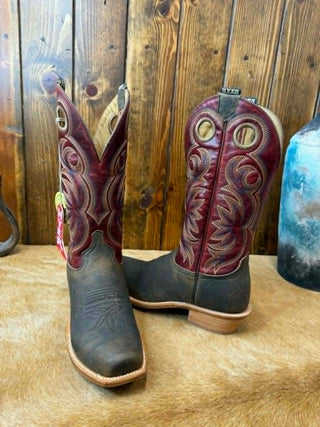 A pair of cowboy boots with dark brown leather bodies and burgundy shafts featuring intricate western embroidery, displayed on a tan hide against a rustic wooden plank wall.