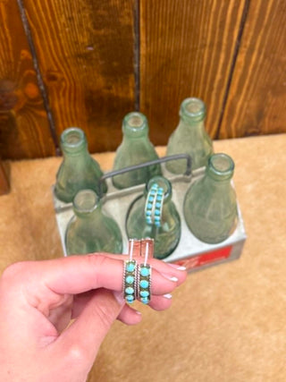 Set of six green glass bottles in a metal carrier with a hand holding turquoise earrings in front.