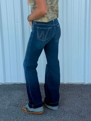 A person wearing dark blue flare jeans with contrast stitching and cuffed hems, standing in front of a white corrugated metal wall.