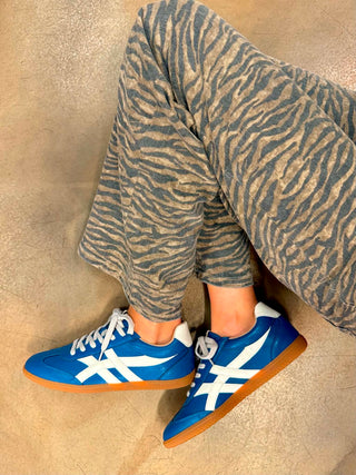 A person wearing blue and white Asics sneakers with zebra-print lounge pants, sitting on a textured beige floor.