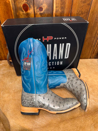 A pair of cowboy boots displayed in front of their black branded box on a brown fur surface against a wooden wall. The boots have bright blue western shafts with carved stitching and gray ostrich-textured feet.
