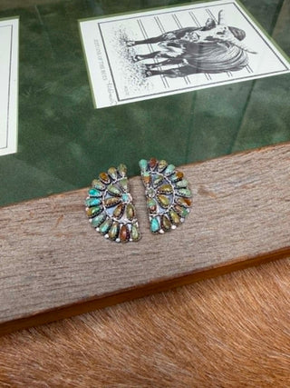 A pair of silver-toned Kachepa-style turquoise and multicolor stone brooches displayed on a wooden block over a fur surface, with a green glass case and an illustration card in the background.