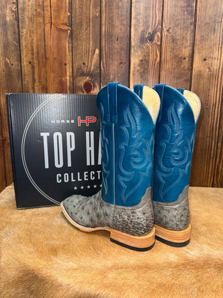 A pair of blue and gray western cowboy boots displayed next to their black 'Top Hat Collect' shoebox against a rustic wooden plank wall and tan hide background.