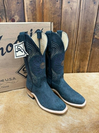 A pair of dark teal western cowboy boots with floral embroidery and light tan leather trim, displayed next to their cardboard box on a tan hide surface against a rustic wooden plank background.