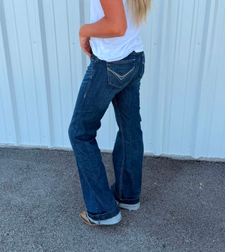 A person wearing dark blue flare jeans with contrast stitching, a white t-shirt, and white sneakers, standing in front of a white corrugated metal wall.