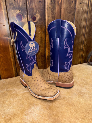 A pair of cowboy boots with blue shafts and tan ostrich-textured feet, displayed on a tan faux fur rug against a rustic wooden plank wall. A round blue tag on the left boot reads 'Originals'.