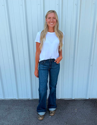 A woman with long blonde hair stands in front of a white corrugated metal wall, wearing a white short-sleeve t-shirt, blue flare jeans, and brown shoes.
