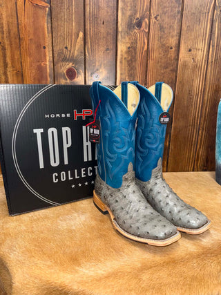 A pair of HP Horned Horse cowboy boots with bright blue shafts and gray ostrich-textured feet, displayed next to a black 'Horse Top Hat Collection' box on a brown hide surface against a wooden plank wall.