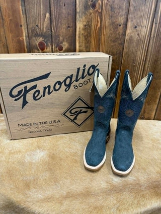 A pair of blue suede Fenoglio cowboy boots standing next to their original wooden box on a tan faux fur surface against a rustic wood plank background.