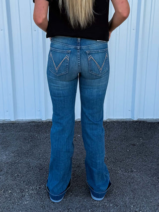 A person viewed from behind wearing blue flare jeans with contrast stitching and a black top, standing outdoors against a light blue corrugated metal wall.